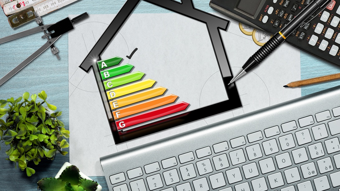 Ett professionellt skrivbord med en datorskärm som visar ett energiklassificeringsdiagram överlagrat på en hussiluett. A-G energiklassificeringsskalan använder färgkodning från grönt (mest effektivt) till rött (minst effektivt), omgiven av ritverktyg och en miniräknare, vilket indikerar arbete med energibedömning för byggnader.